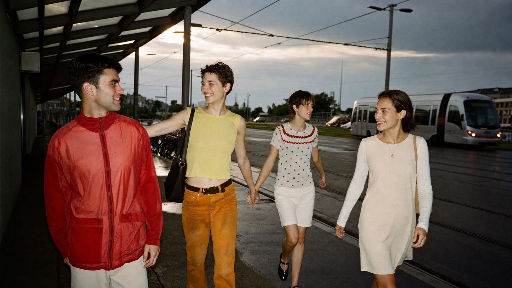 Four friends wearing red, yellow and white walk together toward the camera at a tram stop after sunset.