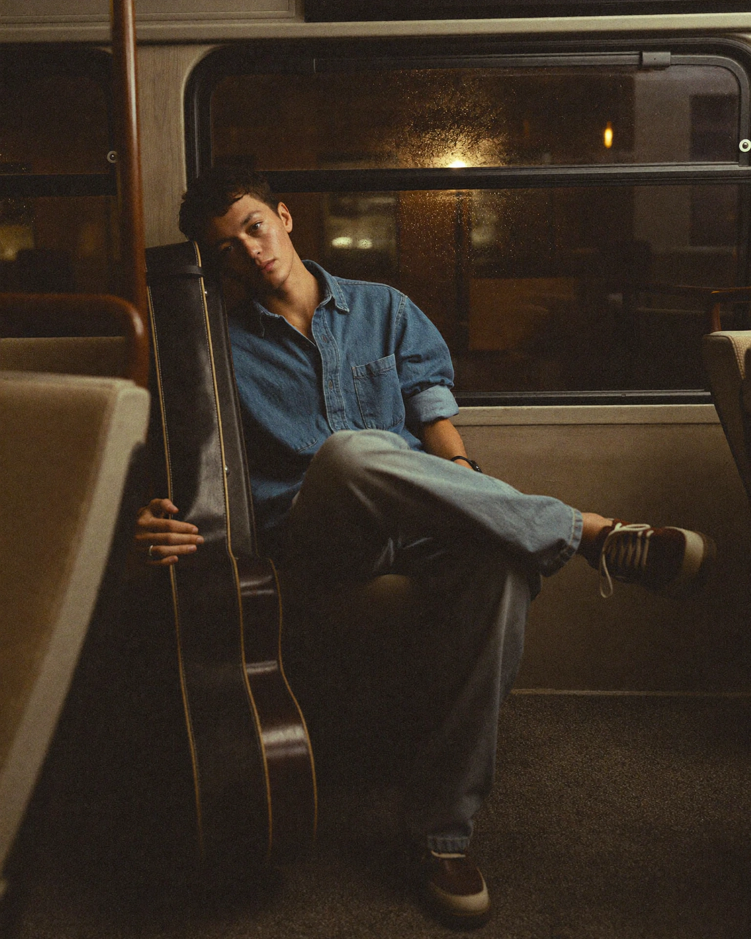 A young man in a denim shirt and jeans sits on a night bus beside a guitar case and a rain-speckled window.