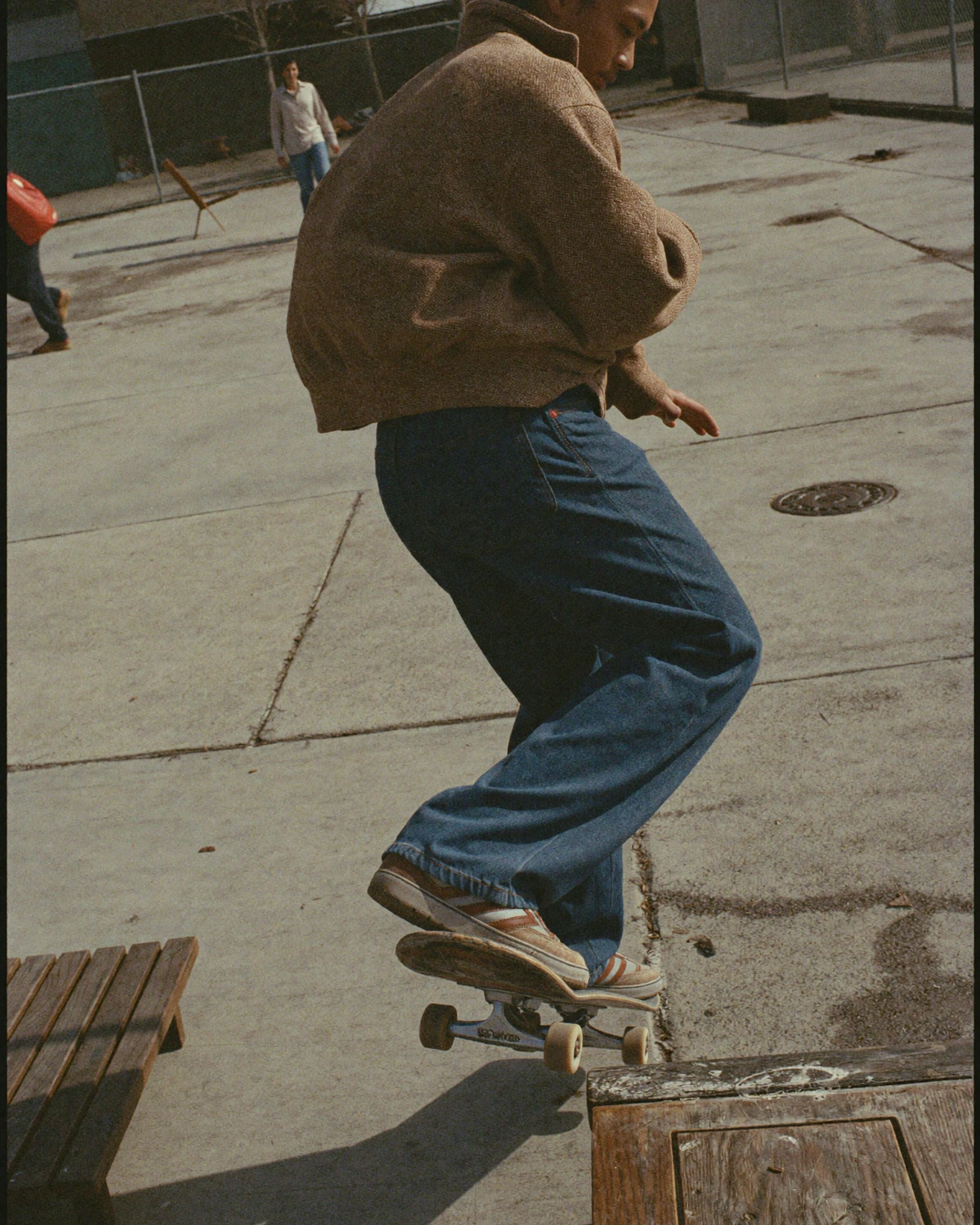 A skateboarder in loose blue jeans and a brown jacket rides beside a wooden box on a sunlit concrete lot.