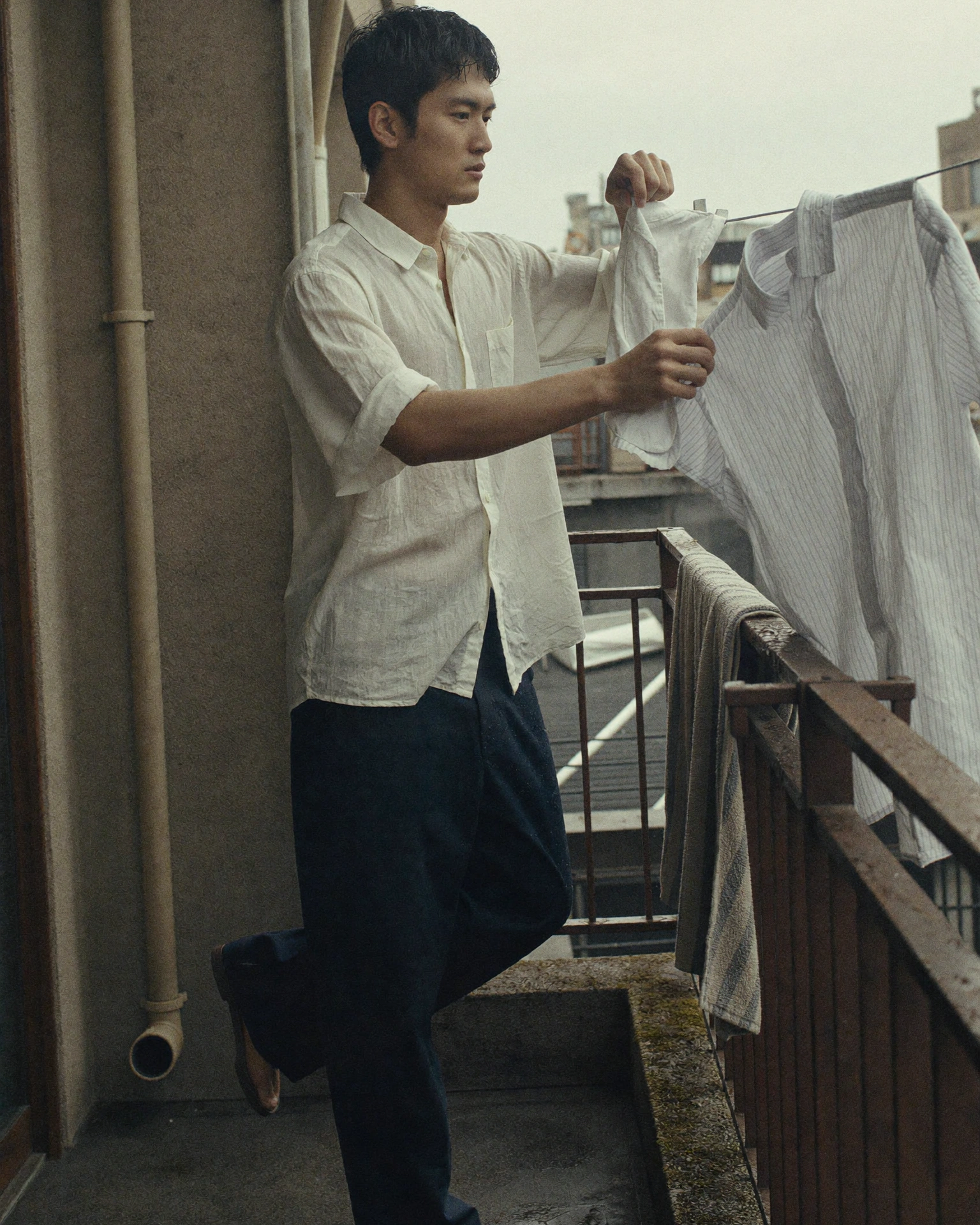 A man clips a white shirt to a laundry rail on a narrow concrete balcony.