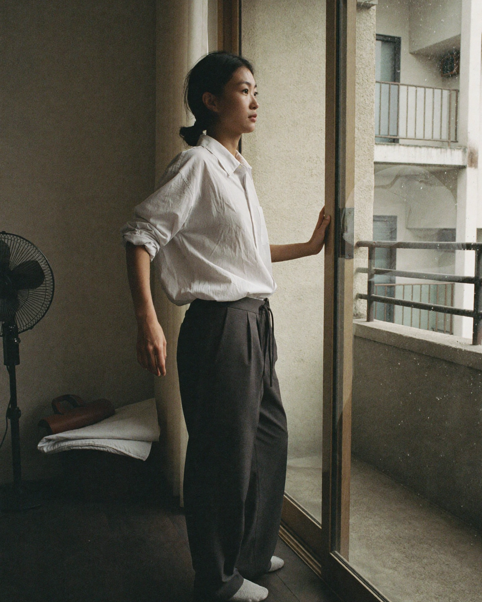 A woman in a white shirt and dark trousers stands beside an open sliding balcony door in a spare apartment.