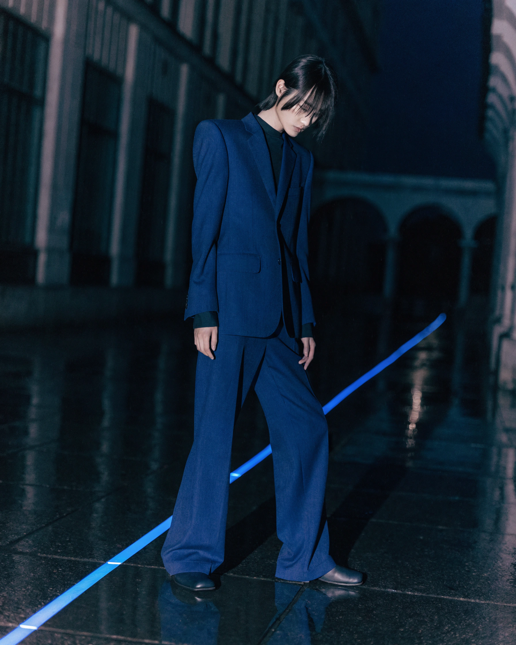 A tailored figure and one diagonal light repeat across rain-polished pavement.