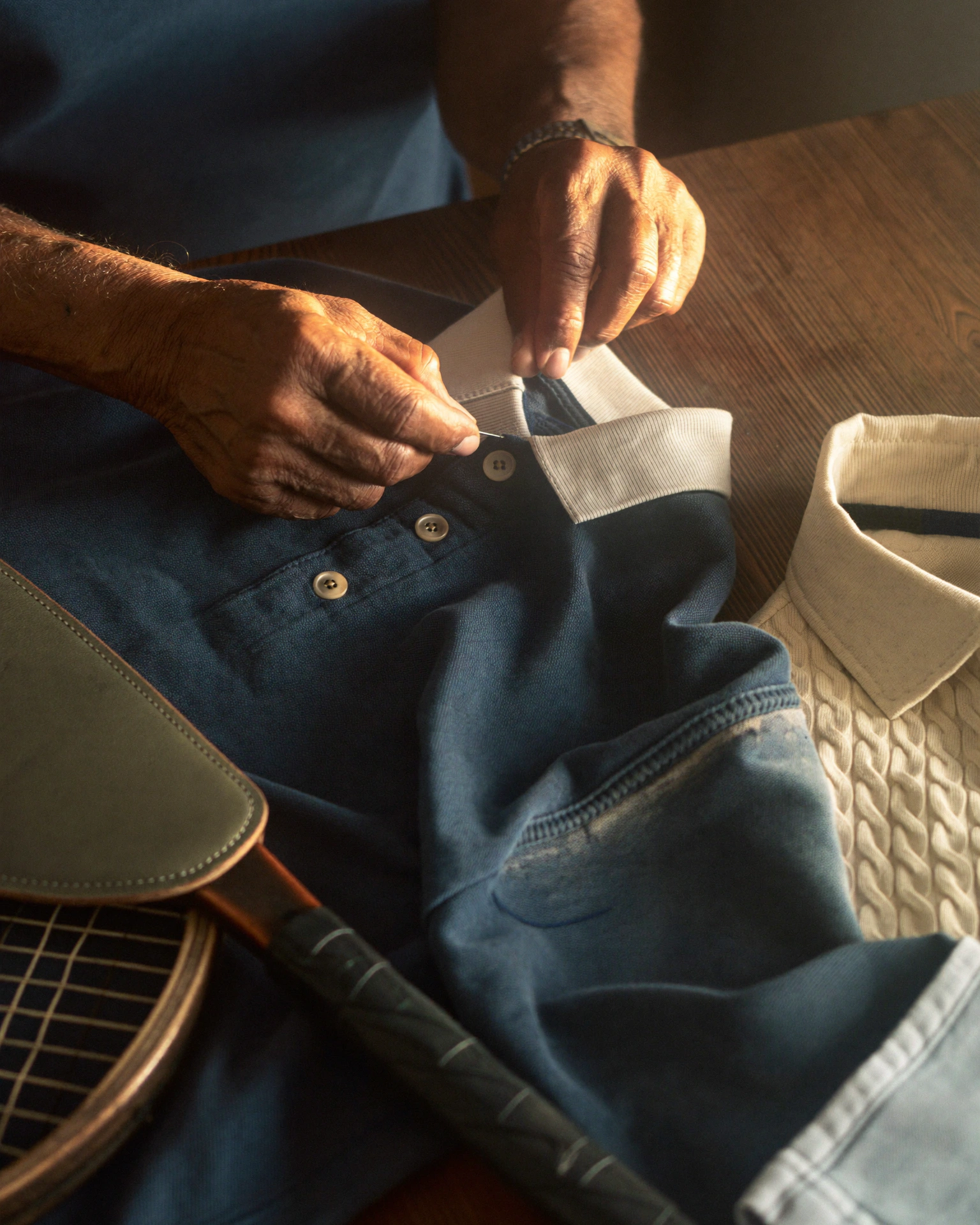 Hands work at the collar of a navy Polo-style shirt beside cream cable-knit fabric and a wooden racquet.