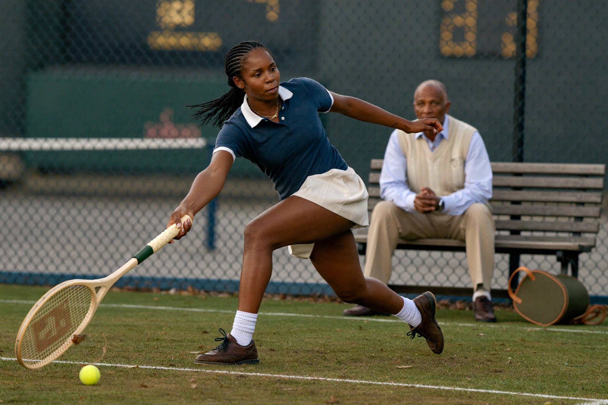 A woman in navy and white lunges for a low tennis ball while an older man watches from a bench.