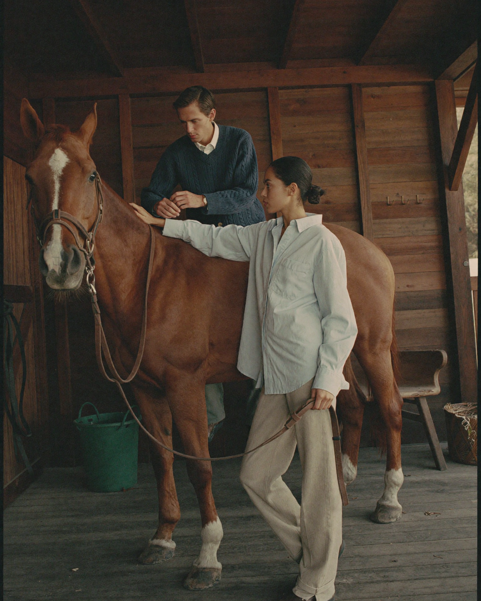 A woman holds reins beside a chestnut horse while a man in a navy sweater leans over its back inside a wooden stable.