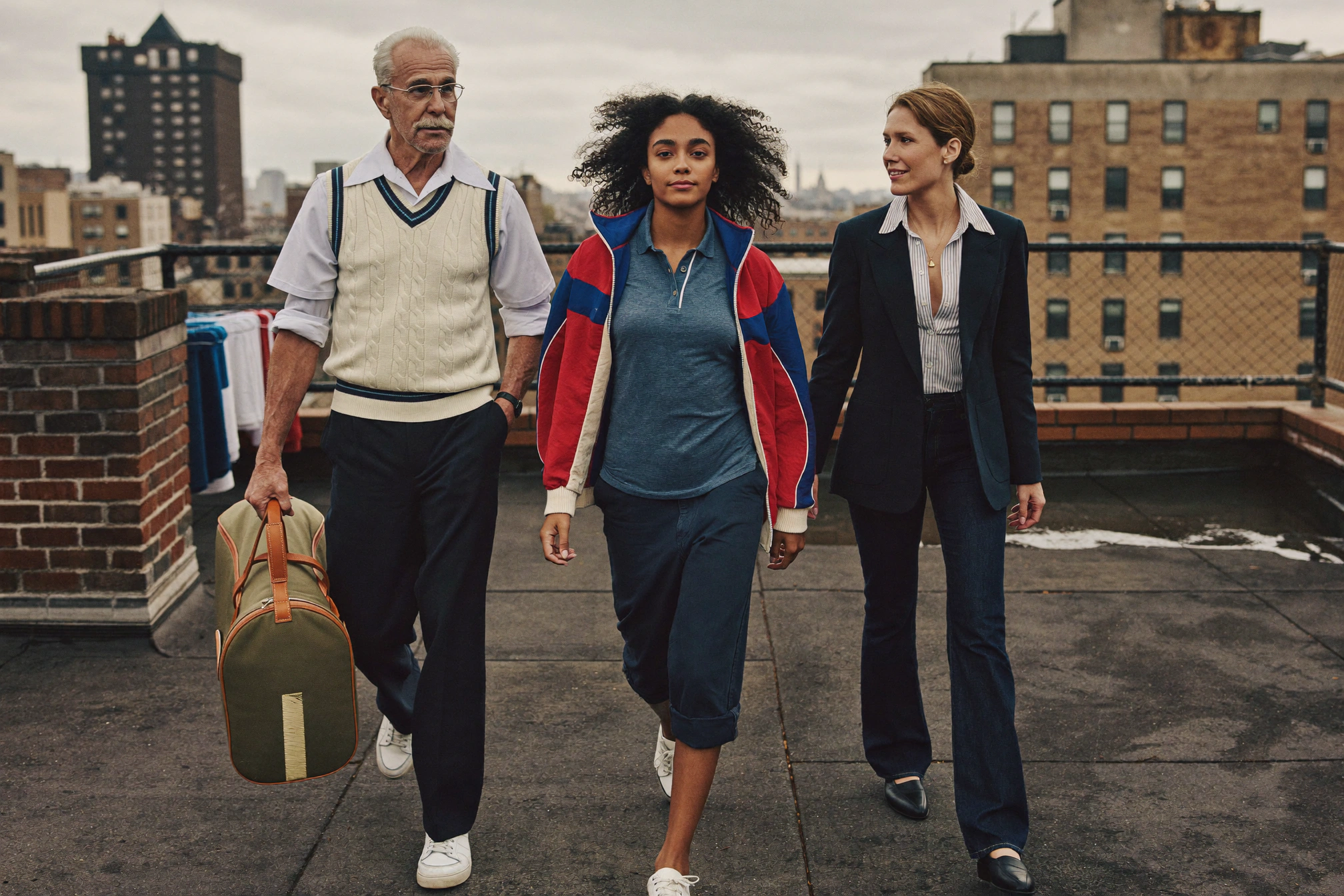 An older man and two women cross a rooftop in layered navy, cream, red, and blue clothing while carrying a racquet bag.