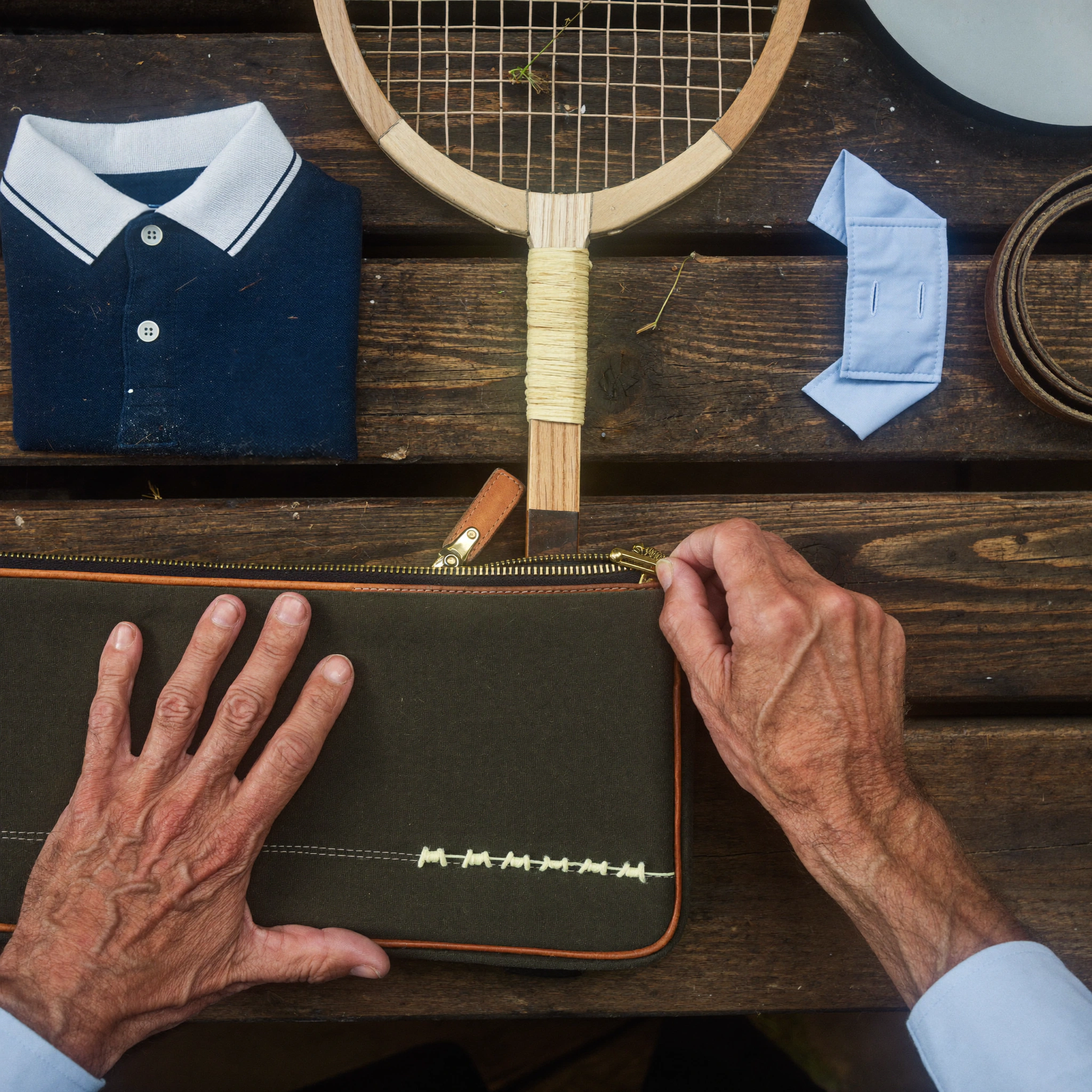 A top-down view shows hands above a folded navy polo shirt, a wooden racquet, and a small green case.