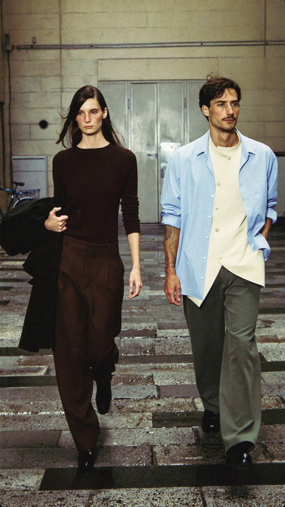 A woman in brown and a man in a pale blue jacket descend wet stairs outside an industrial building.
