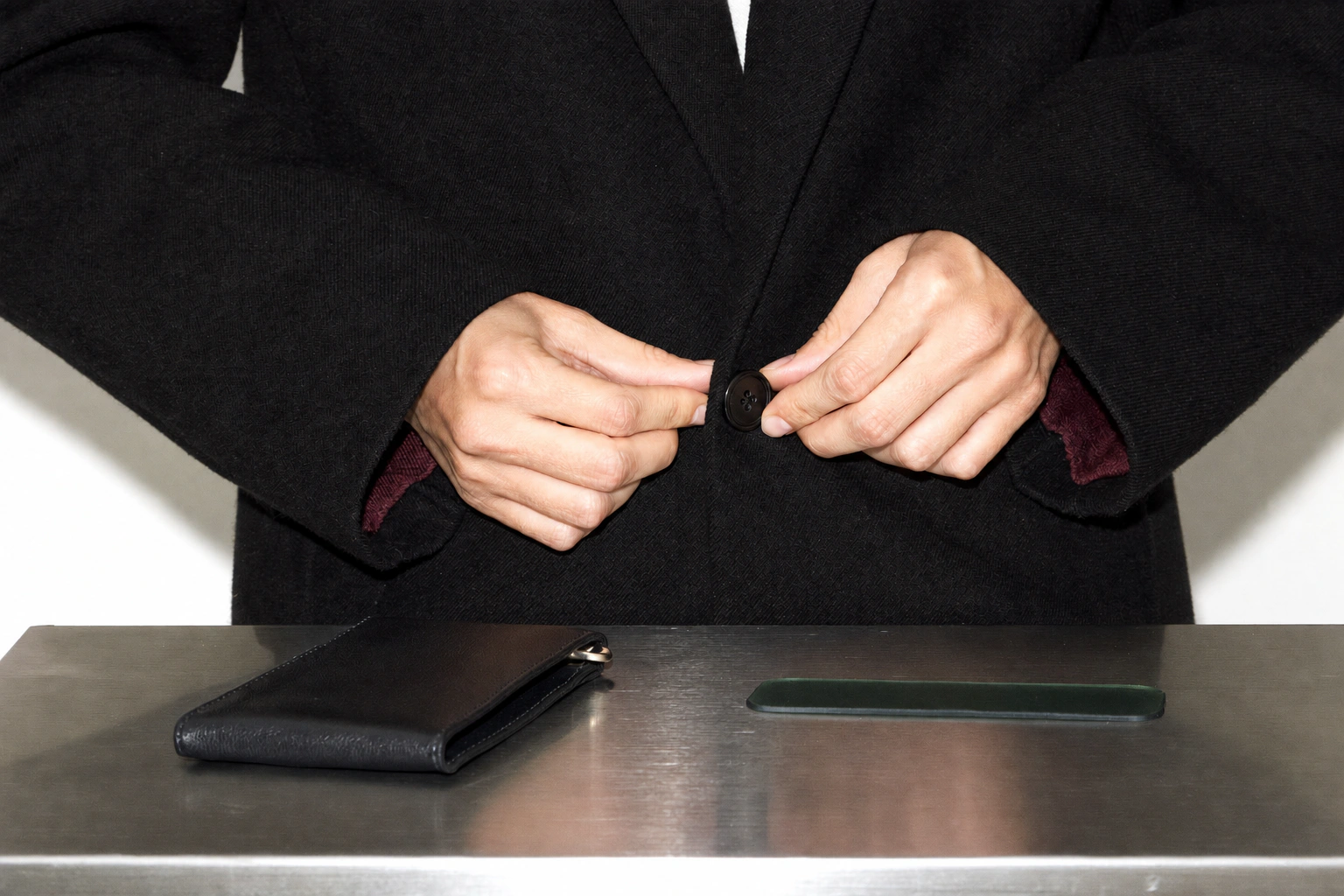 Hands fasten a black coat beside a metal wallet and a narrow strip of green material.