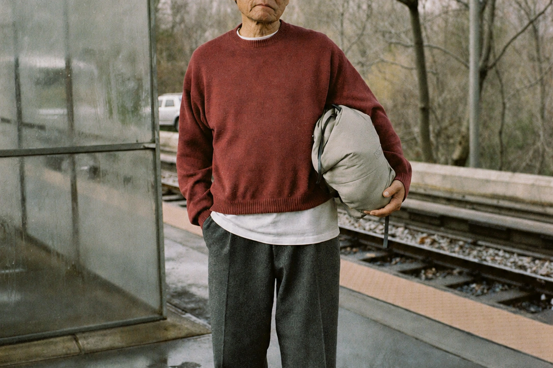 Close view of an older commuter's red sweater and grey trousers, carrying a light padded layer beside a railway platform.