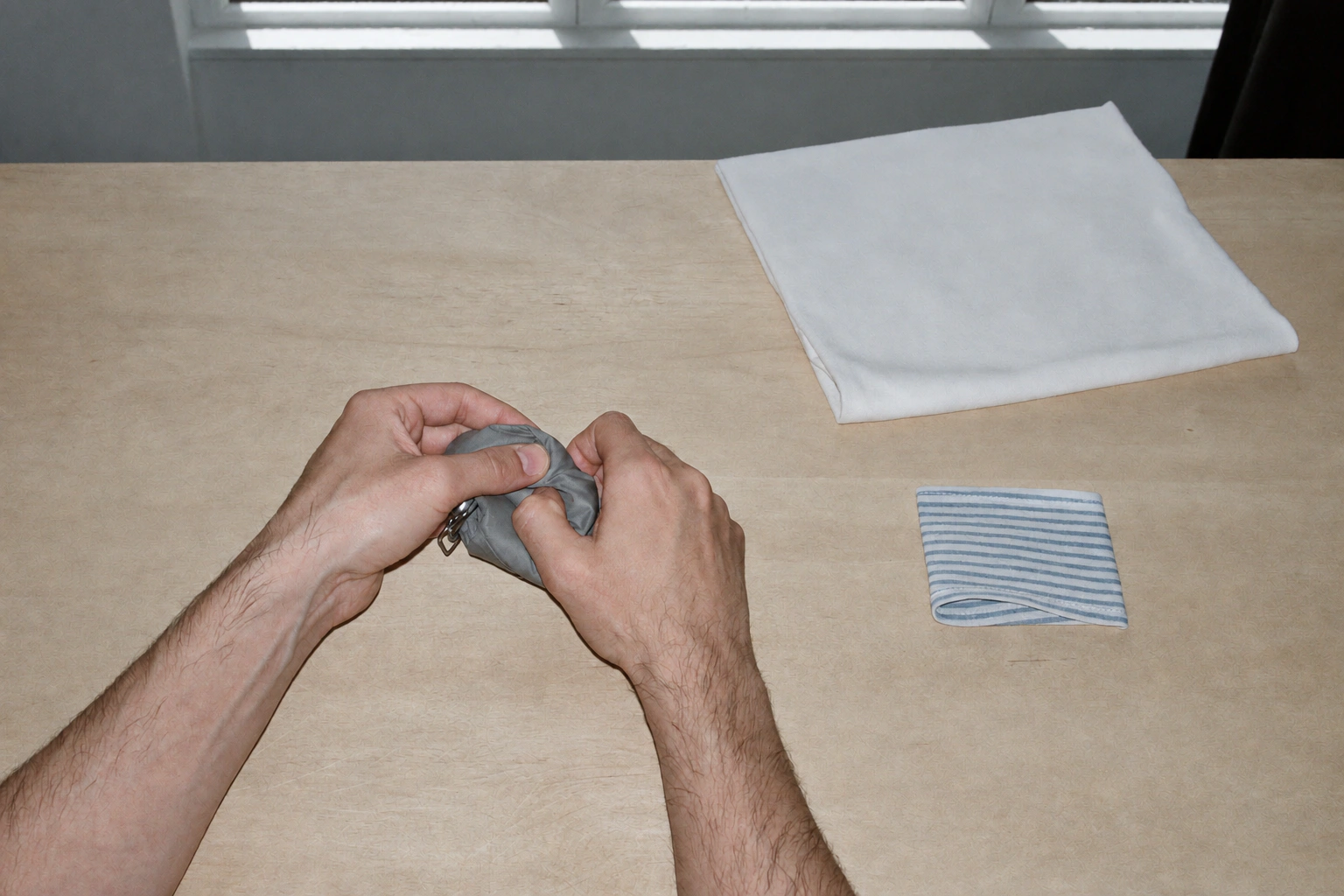 Hands compress a small grey fabric layer beside neatly folded white and striped cloth on a light wooden table.