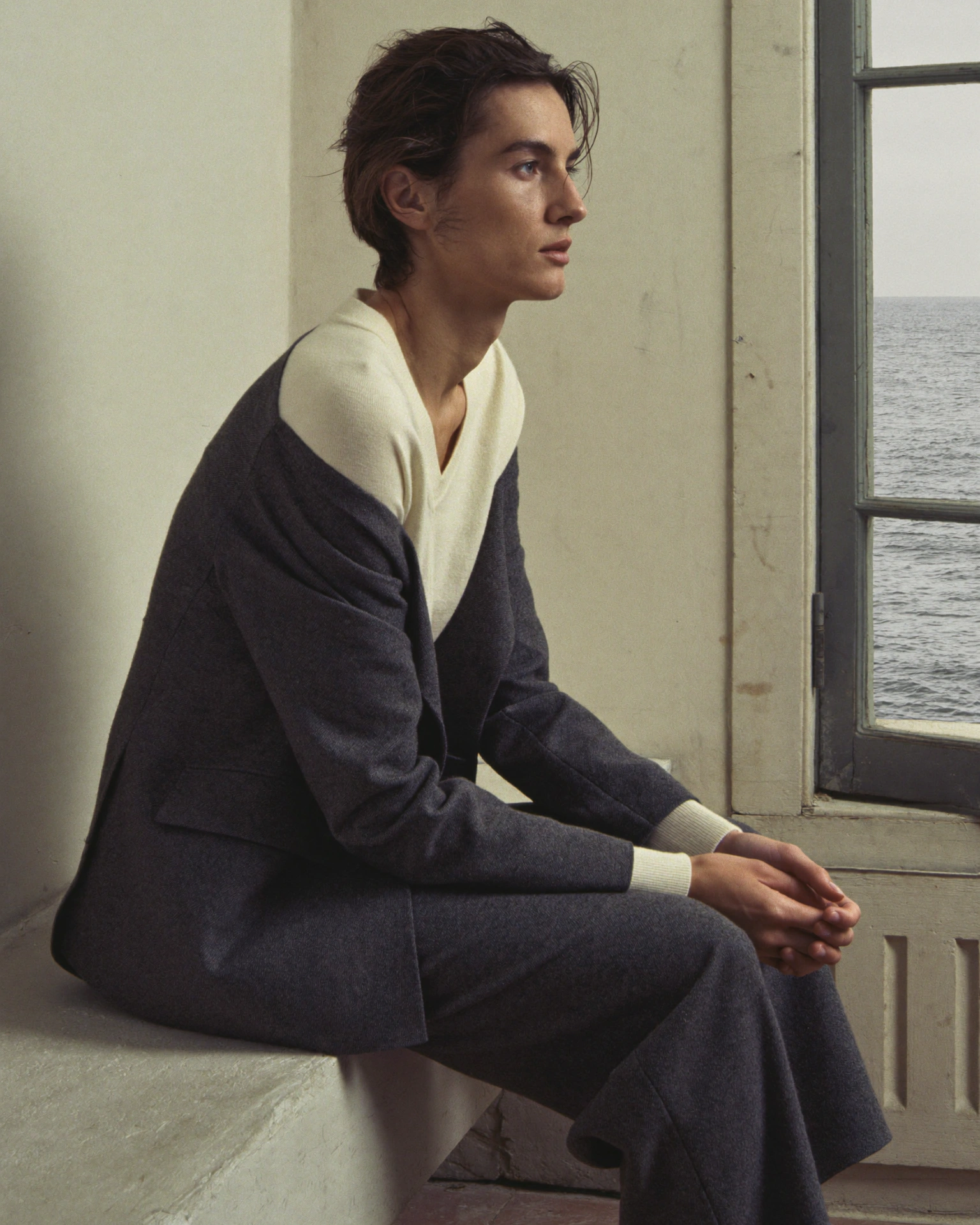 A person in a grey suit and cream knit sits beside a window overlooking the sea.