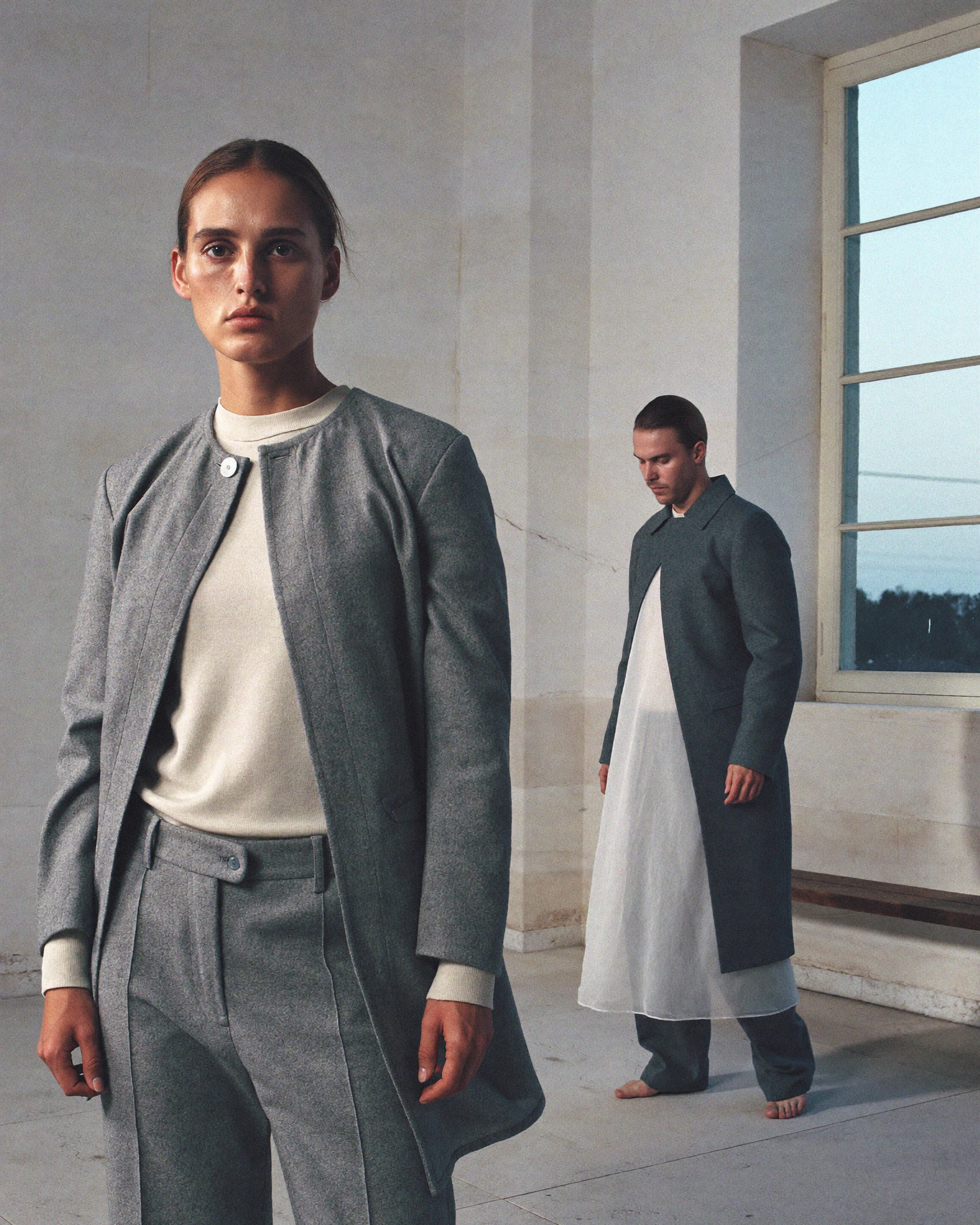 Two models wear grey and cream tailoring in a pale room, one close to the camera and one beside a tall window.